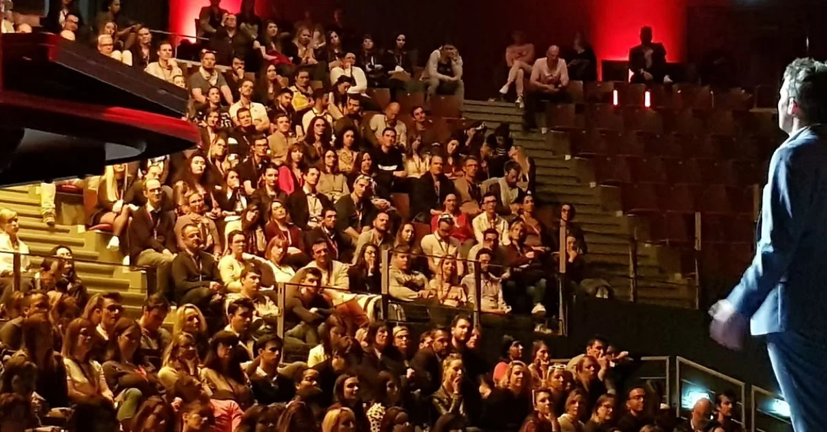 Public attentif dans un amphithéâtre face au conférencier qui entre en scène